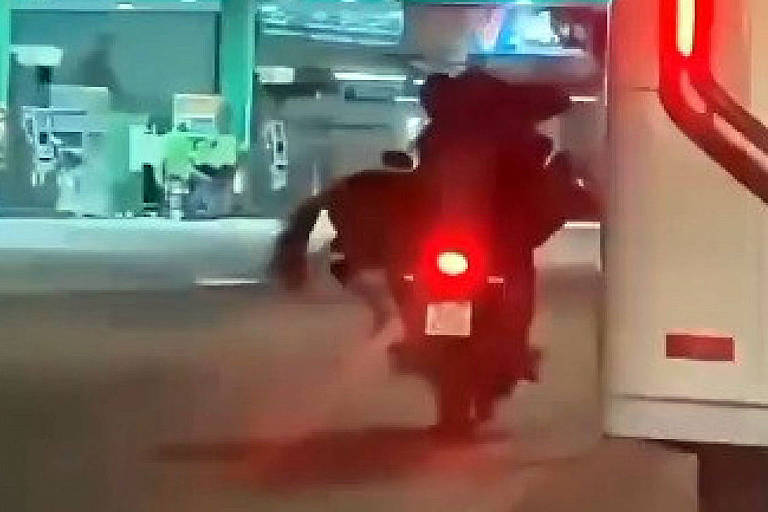 A imagem mostra uma motocicleta em movimento, com um farol traseiro aceso. Ao fundo, há uma estrutura que parece ser uma estação de pedágio ou um posto de gasolina, com várias máquinas visíveis. A cena é noturna, e a iluminação é predominantemente artificial.