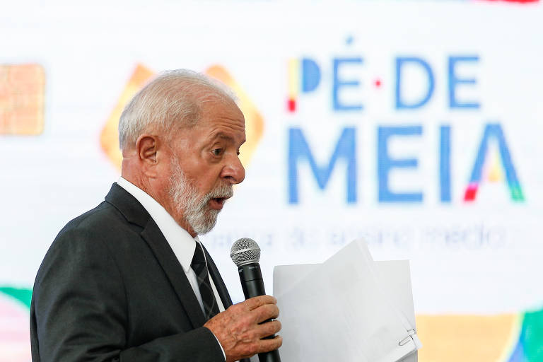 Presidente Lula， de cabelo grisalho e barba， vestido com um terno escuro， está falando ao microfone enquanto segura um papel. Ao fundo， há um painel colorido com as palavras 039;PÉ DE MEIA039; em destaque， com um design gráfico vibrante.