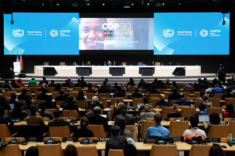 A imagem mostra uma grande sala de conferências com várias pessoas sentadas em cadeiras. No fundo, há um palco com uma mesa retangular e várias cadeiras. Em uma tela grande, há uma apresentação com a imagem de uma pessoa sorrindo e o logotipo da ONU. O ambiente é bem iluminado e organizado, com bandeiras visíveis ao lado do palco.