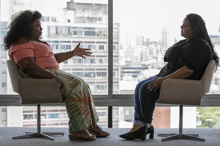 A imagem mostra duas mulheres sentadas em cadeiras em um ambiente interno com grandes janelas ao fundo, que revelam uma vista urbana. A mulher à esquerda tem cabelo cacheado e usa uma blusa rosa e uma saia com estampas coloridas, gesticulando com a mão. A mulher à direita tem cabelo liso e usa uma blusa preta e jeans, ouvindo atentamente. Ambas parecem estar em uma conversa significativa.