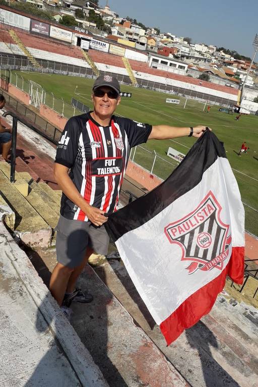 Um torcedor está em um estádio segurando uma bandeira. Ele usa uma camisa listrada em preto, branco e vermelho e um boné. O fundo mostra um campo de futebol e arquibancadas. A bandeira é predominantemente branca com detalhes em preto e vermelho, e possui um emblema no centro.