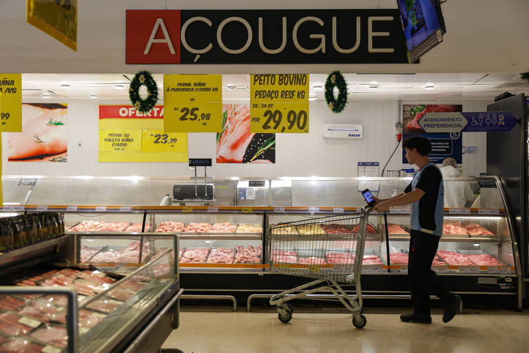 A imagem mostra a seção de açougue de um supermercado. No fundo， há um grande letreiro com a palavra 039;AÇOUGUE039; em destaque. Vários cartazes promocionais estão visíveis， com preços e ofertas. Um funcionário， vestido com uniforme， está empurrando um carrinho ao lado de expositores de carne. A área é bem iluminada e organizada.