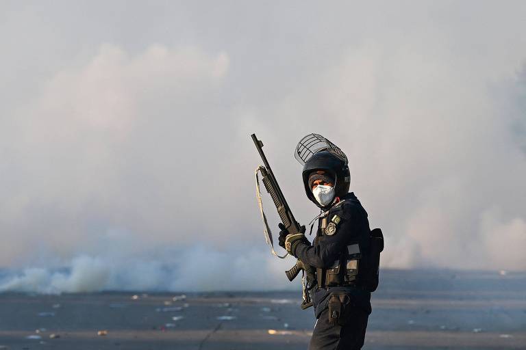 Um policial está em pé, segurando um rifle, em meio a uma cena de conflito. Ele usa um capacete e uma máscara facial, e há fumaça ao fundo, sugerindo uma situação tensa. O ambiente parece caótico, com resíduos visíveis no chão.