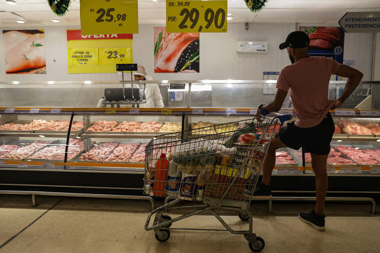 Imagem mostra um consumidor fazendo compras no açougue de um supermercado em São Paulo. Ele está parado com um carrinho cheio de produtos enquanto observa as carnes à venda no local.
