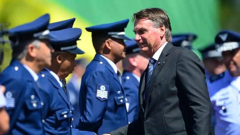 A imagem mostra Bolsonaro, um homem de terno escuro cumprimentando membros das forças armadas, que estão vestidos com uniformes azuis. O fundo apresenta uma bandeira do Brasil, com as cores verde e amarelo visíveis. O homem parece estar em uma cerimônia oficial, interagindo com os militares em uma linha.