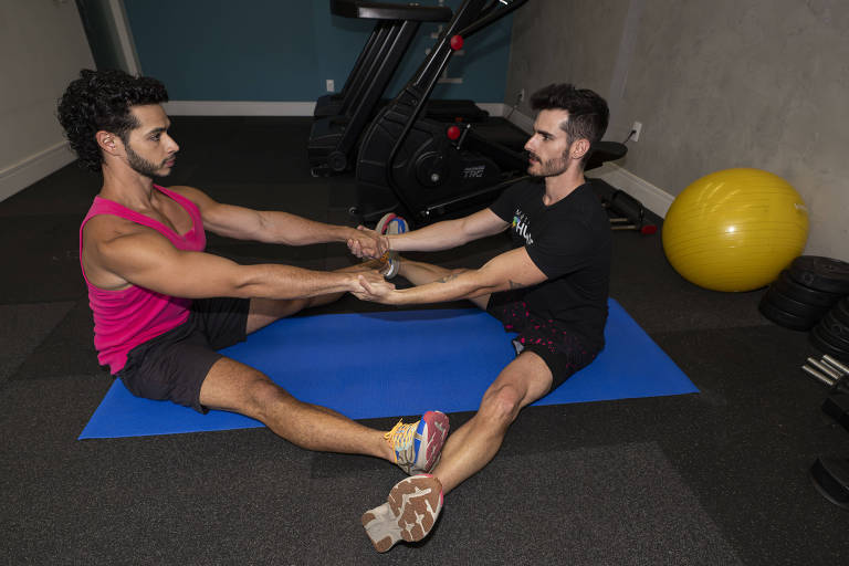 Dois homens jovens， ambos de cabelo escuro e barba estão sentados de frente um para o outro em colchonete azul no chão de uma academia， realizando um exercício de alongamento em dupla. Eles estão segurando as mãos um do outro， com os braços estendidos， e as pernas estão abertas em vê. O homem à esquerda usa uma camiseta rosa e shorts pretos， e o à direita veste camiseta preta e shorts com estampa colorida. Ao fundo， há equipamentos de ginástica e uma bola de exercício amarela.