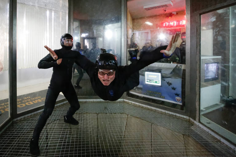 A imagem mostra duas pessoas em um túnel de vento. Uma delas está flutuando no ar, com os braços abertos e uma expressão de alegria, enquanto a outra está em pé, observando. Ambas estão vestidas com trajes pretos e capacetes. O ambiente é iluminado e há uma tela visível ao fundo.