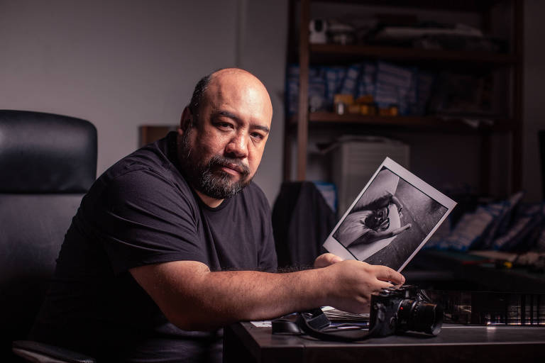 Um homem com cabelo curto e barba está sentado diante de uma mesa， segurando uma impressão em preto e branco de uma fotografia. Ele usa uma camiseta preta e parece estar em um ambiente de trabalho， com uma câmera e outros materiais fotográficos sobre a mesa. Ao fundo， há prateleiras com objetos e materiais organizados.