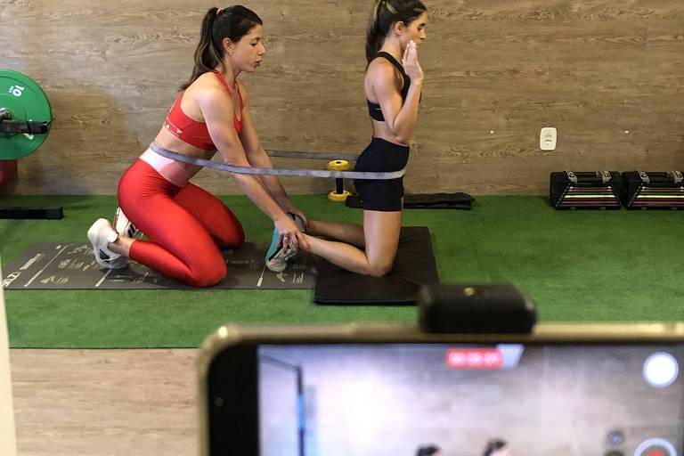 A imagem mostra duas mulheres em um ambiente de treino. A mulher à esquerda， vestindo um conjunto de treino vermelho， está de joelhos e segurando uma faixa elástica， enquanto a mulher à direita， vestindo um top preto， está em pé com a mão no rosto. Um celular em um suporte captura a cena， mostrando a mesma posição das duas mulheres. Ao fundo， há equipamentos de ginástica， como pesos e uma bola de exercício