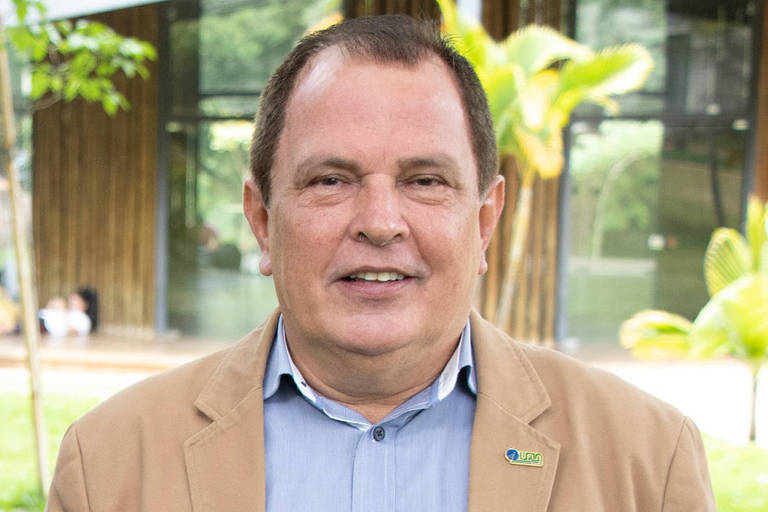 Um homem sorridente está posando para a foto. Ele usa um paletó bege e uma camisa azul clara. Ao fundo， há um ambiente natural com plantas e uma construção moderna. A imagem transmite uma sensação de tranquilidade e profissionalismo.