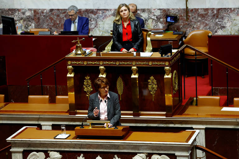 A imagem mostra uma sessão do parlamento, com uma mulher em pé falando ao microfone em um púlpito na frente. Ao fundo, outra mulher observa sentada em uma mesa elevada, enquanto um homem está ao fundo, em uma mesa com um computador. O ambiente é decorado com painéis de mármore e há cadeiras e mesas dispostas para os membros do parlamento.