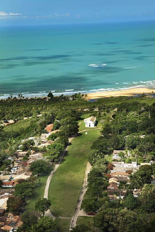 Vista do campo aberto do Quadrado， cercado por casas que hoje são lojas， restaurantes e pousada， com uma igrejinha ao fundo e a praia atrás