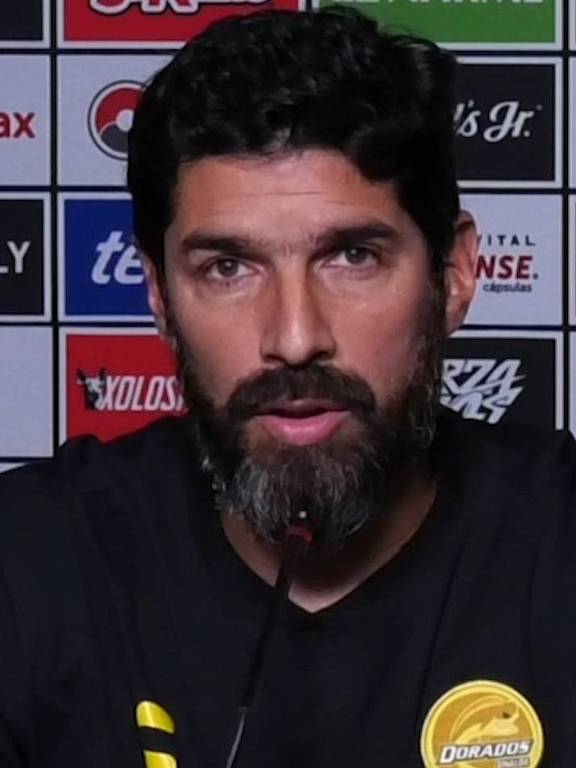 Um homem com barba e cabelo escuro, vestindo uma camiseta preta com um logo amarelo, está sentado em uma mesa de coletiva de imprensa. Ele fala em um microfone, com as mãos apoiadas na mesa. Ao fundo, há uma parede com vários logotipos de patrocinadores. Abaixo dele, está escrito: 'pero que nuestro corazón'.