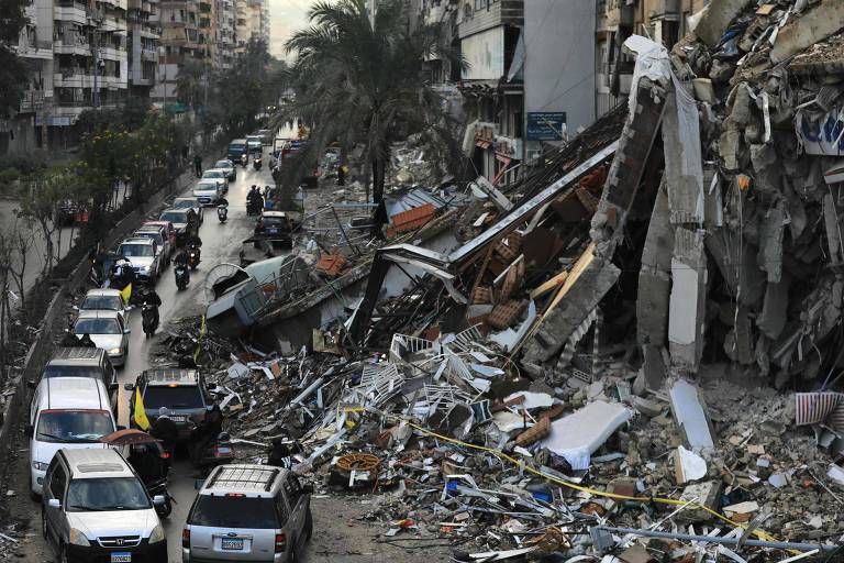 A imagem mostra uma área urbana devastada, com prédios parcialmente desmoronados e escombros espalhados pela rua. Vários carros estão estacionados ao longo da via, e algumas pessoas estão visíveis, possivelmente avaliando os danos. Há árvores ao fundo, e a cena transmite um sentimento de destruição e desolação.