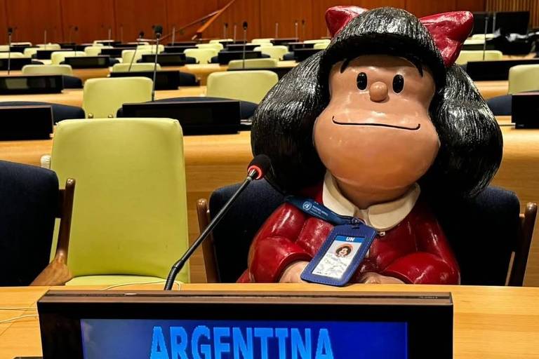 A imagem mostra uma figura de Mafalda, um personagem de quadrinhos, sentada em uma mesa de conferência da ONU. Ela está vestida com um vestido vermelho e uma fita na cabeça, com um crachá visível que indica sua afiliação à ONU. À sua frente, há uma placa com a palavra 'ARGENTINA' em letras azuis. O ambiente ao fundo é uma sala de conferências com várias cadeiras e mesas dispostas.