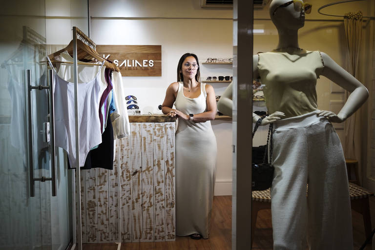 A imagem mostra o interior de uma loja de roupas. Uma mulher está em pé atrás de um balcão de madeira, usando um vestido longo e óculos. Ao lado dela, há um manequim vestido com uma blusa e calças brancas. No fundo, há prateleiras com óculos de sol e roupas penduradas em cabides. A iluminação é suave e o ambiente é acolhedor.