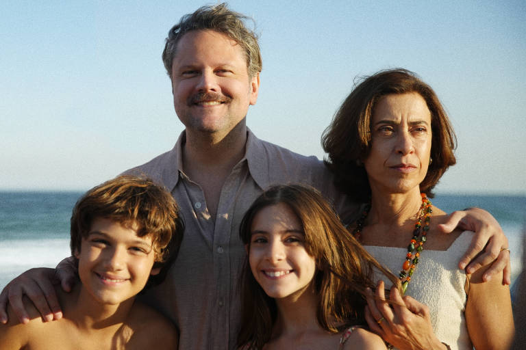  A imagem mostra uma família posando na praia. Um homem com bigode e camisa clara está no centro， sorrindo. À sua esquerda， um menino com cabelo castanho e ondulado sorri. À sua direita， uma mulher com cabelo castanho e liso， usando um colar colorido， tem uma expressão séria. Uma menina com cabelo longo e castanho， também sorrindo， está à frente do homem. O fundo apresenta o mar e o céu claro.