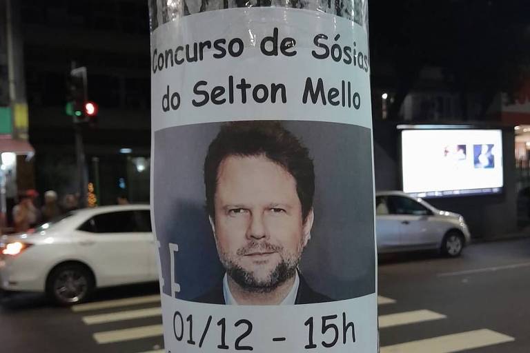 Um cartaz colado em um tronco de árvore anuncia um concurso de sósias do Selton Mello. O cartaz contém uma foto do ator e informações sobre o evento, incluindo a data (01/12), horário (15h) e local (Saguão do Estação NET Rio). Também menciona que o prêmio é de 5 cortesias para qualquer filme