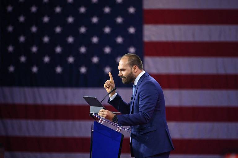 Foto mostra Eduardo Bolsonaro de perfil falando em um palanque com a bandeira dos Estados unidos ao fundo. Ele é um homem branco com barba curta, calvície. Bolsonaro está com dedo em riste e veste um terno azul-escuro.