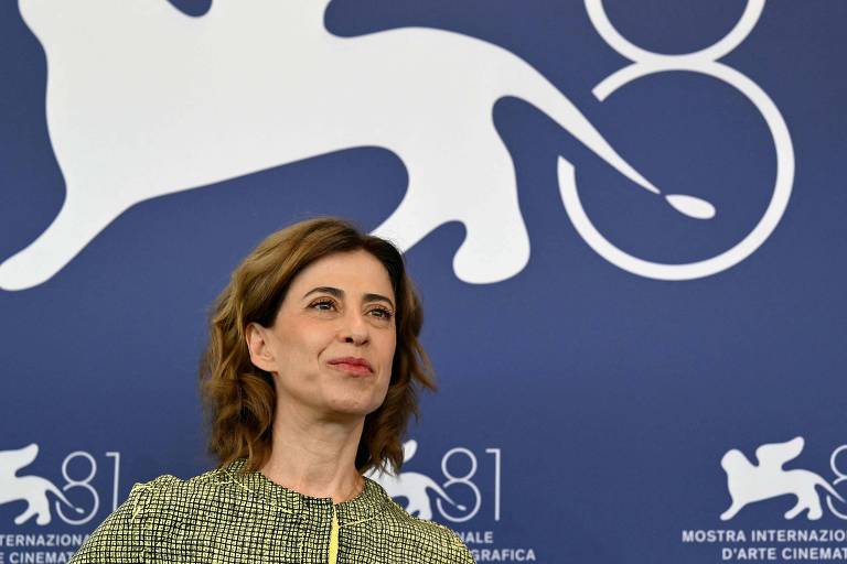 Fotografia mostra a atriz brasileira Fernanda Torres. Ela é branca, usa um vestido verde, tem cabelo castanho claro e está na frente de um painel azul-marinho.