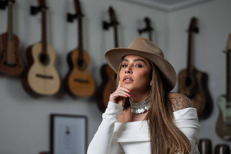 A imagem mostra uma mulher com cabelo longo e liso, usando um chapéu claro e uma blusa branca. Ela está posando com a mão no queixo, olhando para a câmera. Ao fundo, há várias violões penduradas na parede. A mulher usa um colar de corrente e brincos, e tem uma tatuagem visível no braço