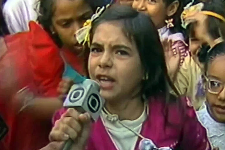 A imagem mostra um grupo de crianças em uma entrevista. Uma menina em destaque, com cabelo escuro e uma blusa rosa, está falando para um microfone. Ela parece estar expressando uma opinião forte. Ao fundo, outras crianças estão visíveis, algumas com acessórios no cabelo, como laços e tiaras. A cena é animada e cheia de energia.