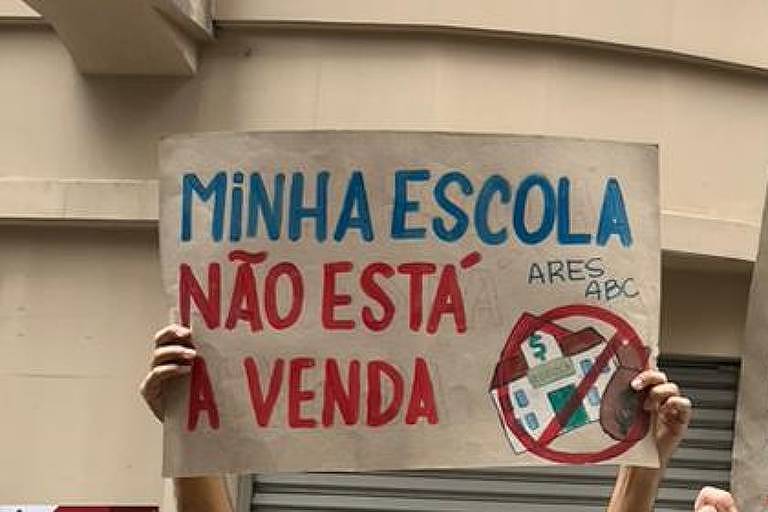 A imagem mostra um protesto em que duas pessoas seguram cartazes. O primeiro cartaz diz 039;MINHA ESCOLA NÃO ESTÁ A VENDA039; e o segundo diz 039;TARCÍSIO LADRÃO TIRA A MÃO DA MINHA ESCOLA039;. Ao fundo， há um edifício com janelas grandes e uma placa de 039;ALUGA039;. O ambiente é urbano， com várias pessoas ao redor.