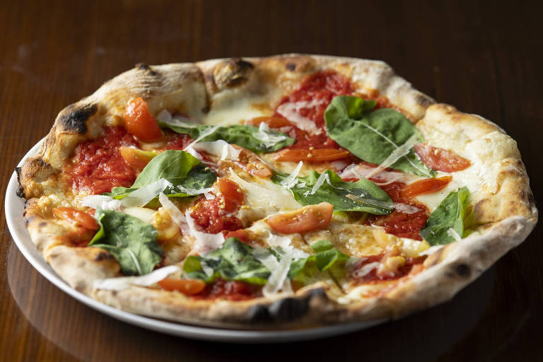 A imagem mostra uma pizza sobre uma mesa de madeira. Ela está dentro de um prato branco. Há pedaços de folhas verdes que aparentam ser rúcula ou algum vegetal fisicamente similar. O alimento também tem tomate seco e queijo derretido