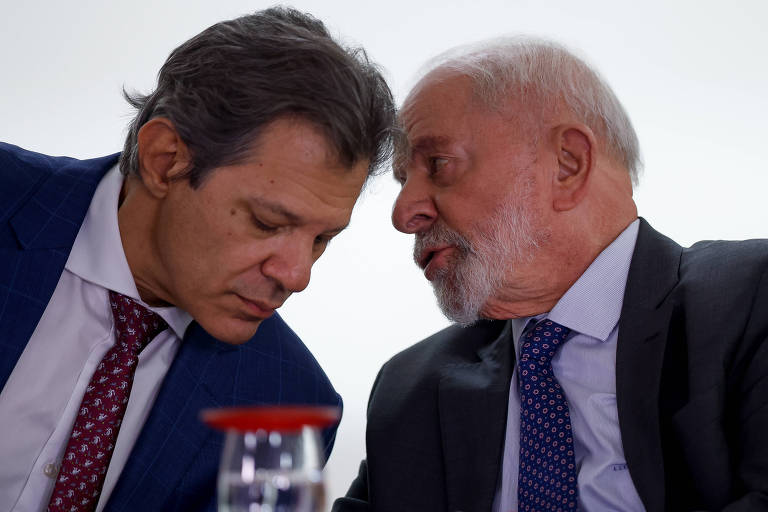 A imagem mostra dois homens em um ambiente formal， inclinando-se um em direção ao outro， aparentemente em uma conversa íntima. O homem à esquerda (Haddad) usa um terno azul com uma gravata vermelha e tem cabelo escuro. O homem à direita (Lula) está vestido com um terno cinza e uma gravata azul， com cabelo grisalho. Ambos parecem concentrados na conversa.