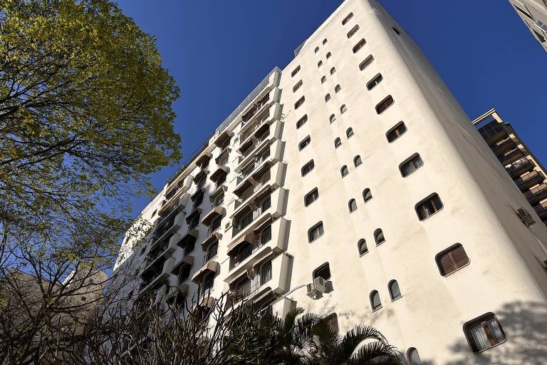 A imagem mostra um edifício residencial de cor branca, com várias janelas retangulares. A foto é tirada de baixo para cima, destacando a altura do prédio. No primeiro plano, há árvores com folhas verdes, e o céu está limpo e azul ao fundo.