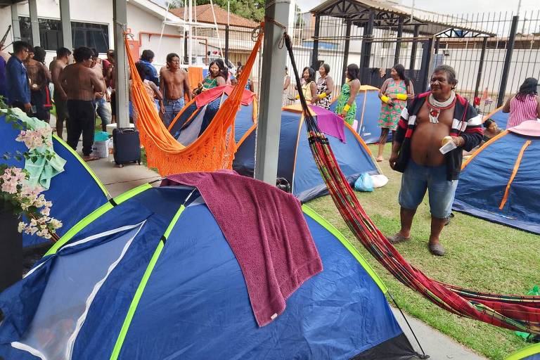 A imagem mostra um grupo de pessoas em um espaço coberto, com várias barracas de camping azuis e redes penduradas. Algumas pessoas estão sem camisa, interagindo entre si, enquanto outras estão sentadas ou em pé. O ambiente parece ser um acampamento, com um gramado ao fundo e uma estrutura de telhado. Há também uma mala preta e flores em um vaso próximo às barracas.