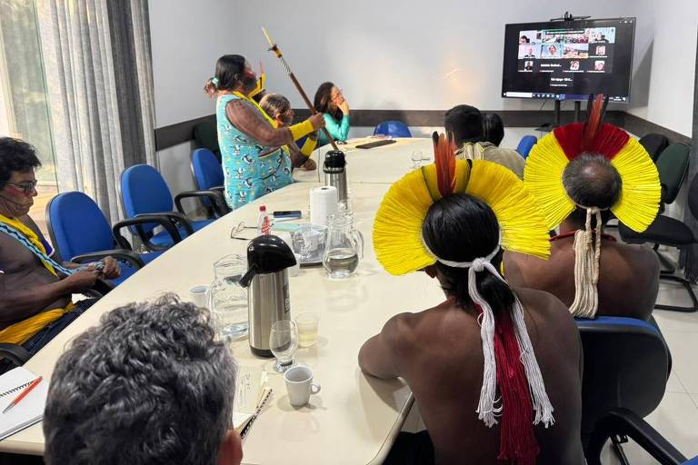 A imagem mostra uma reunião em uma sala com um grupo de pessoas, incluindo representantes indígenas. Alguns participantes estão usando cocares coloridos, predominantemente amarelos e vermelhos. Uma mulher em pé, vestindo uma roupa azul com estampas, está segurando um bastão. Na mesa, há garrafas de água, copos e cadernos. Ao fundo, uma tela exibe uma videoconferência com outras pessoas. A sala tem janelas com cortinas e cadeiras azuis.