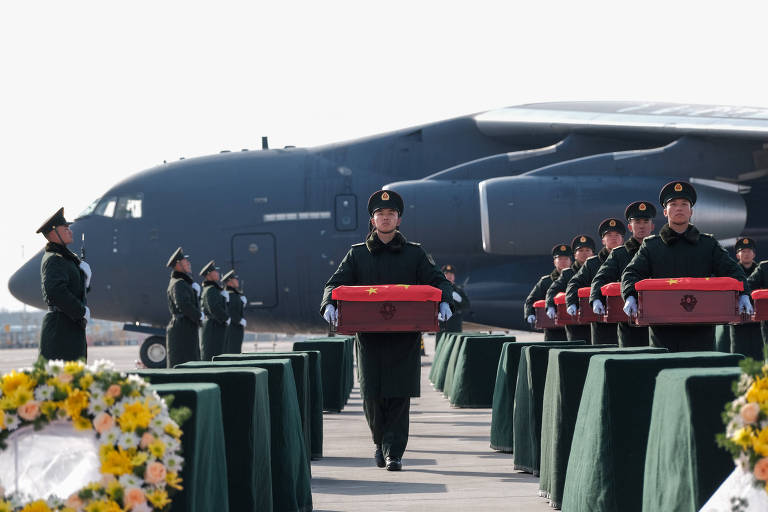 A imagem mostra uma cerimônia militar em um aeroporto, onde soldados em uniformes carregam caixões vermelhos. Ao fundo, um grande avião militar está estacionado. Há mesas cobertas com toalhas verdes e arranjos de flores em primeiro plano. Os soldados estão alinhados em formação, prestando homenagem.