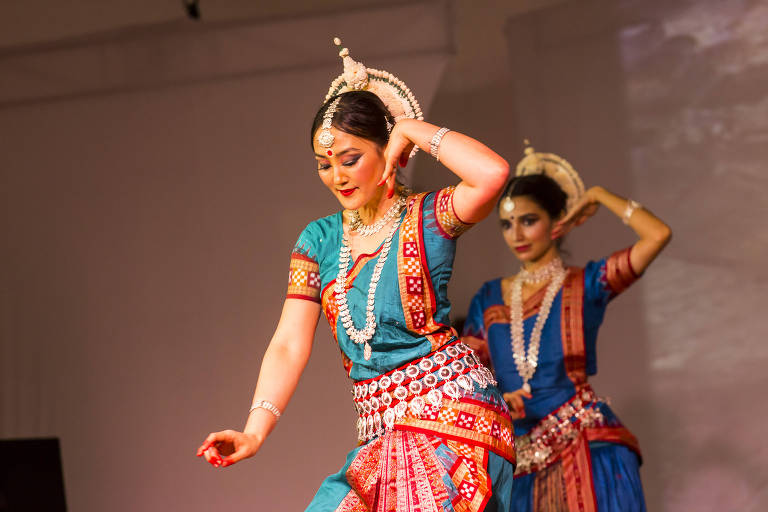 Duas dançarinas em trajes tradicionais estão se apresentando em um palco. A dançarina da frente está em movimento, com um sorriso suave e uma mão levantada, enquanto a dançarina ao fundo observa. Ambas usam roupas coloridas, adornadas com joias e detalhes elaborados. O fundo é neutro, destacando as dançarinas.