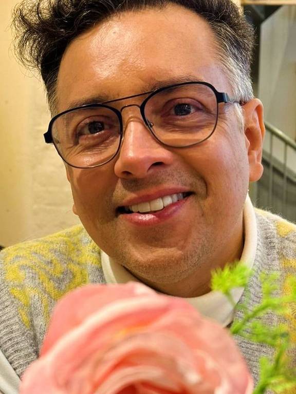 Um homem sorridente está sentado em uma mesa, com um suéter cinza e amarelo. Ele usa óculos e tem cabelo encaracolado. À sua frente, há uma rosa cor-de-rosa em destaque, com folhas verdes ao fundo. O ambiente parece ser um café ou restaurante, com uma decoração simples ao fundo.