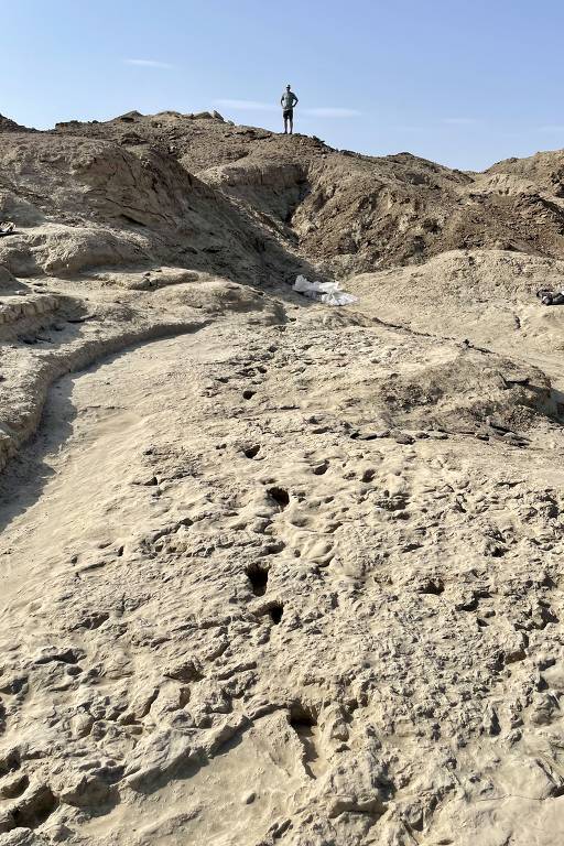 A imagem mostra um terreno árido e irregular, com formações de solo seco e marcas de pegadas. No fundo, uma figura humana está em pé no topo de uma elevação, observando a paisagem. O céu está claro, com algumas nuvens.