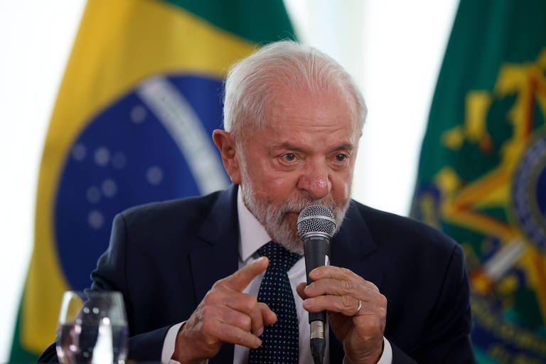 Um homem de cabelo grisalho e barba branca está falando em um microfone. Ele usa um terno escuro e gesticula com a mão. Ao fundo, estão as bandeiras do Brasil e do governo. Há um copo de água em uma mesa à sua frente.