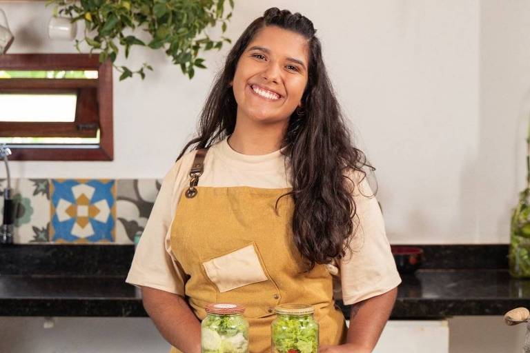 Uma mulher sorridente está em uma cozinha, usando um avental amarelo sobre uma camiseta clara. Ela está em frente a uma mesa de madeira com vários recipientes transparentes contendo diferentes pratos preparados. Dois potes de vidro com saladas estão ao centro. Ao fundo, há prateleiras com plantas e utensílios de cozinha.