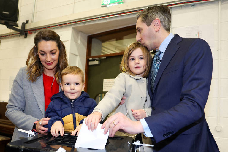 Uma família está participando de uma votação em um local eleitoral. A mãe, com cabelo castanho e vestido vermelho, está ao lado de um menino pequeno, que usa um casaco azul. Uma menina, com cabelo loiro, está ao lado do pai, que está vestido com um terno escuro e gravata. O menino está colocando um papel na urna de votação.