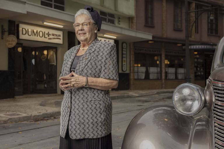 Em foto colorida, mulher posa em frente ao cenário de uma novela de época