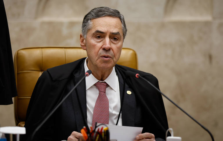 A imagem mostra um homem com cabelo curto e barba， vestindo um terno cinza e uma camisa rosa. Ele está em um ambiente de coletiva de imprensa， cercado por várias pessoas. Ao fundo， é possível ver rostos de mulheres e outras pessoas， algumas segurando microfones e câmeras.