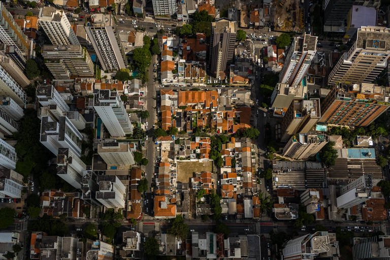 Imagem aérea mostra um quarteirão de casas, com telhados vermelhos, ao centro, cercadas por edifícios altos; a luz solar está concentrada no centro da imagem