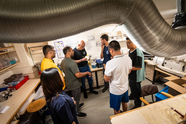 Um grupo de sete pessoas está reunido em um laboratório. Eles estão próximos de uma mesa, onde um homem de camisa preta está escrevendo algo em um papel. Os outros participantes estão atentos, alguns segurando objetos. O ambiente é bem iluminado, com prateleiras ao fundo contendo frascos e materiais diversos. Há um tubo de ventilação visível no teto.