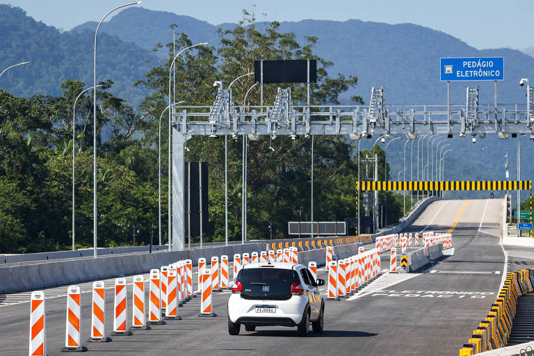 A imagem mostra uma estrada em obras, com um carro branco se aproximando de uma área de pedágio. Há barreiras de segurança laranja e brancas ao longo da pista, e sinais de trânsito visíveis ao fundo, incluindo um sinal azul com a palavra 'Pedágio'. As montanhas são visíveis ao longe, sob um céu claro.