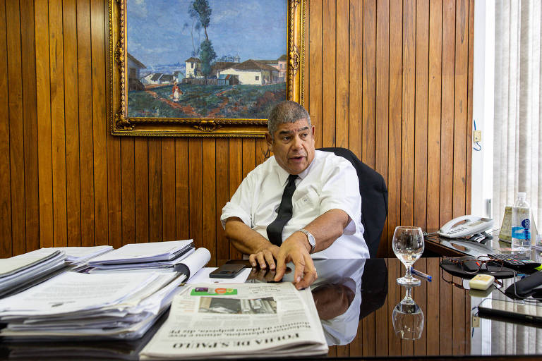 Um homem sentado em uma mesa de escritório， vestindo uma camisa branca e uma gravata preta. Ele está gesticulando com a mão direita enquanto olha para a frente. A mesa está coberta com pastas e um jornal. Ao fundo， há uma pintura na parede e uma janela com cortinas. Um copo de água está sobre a mesa.
