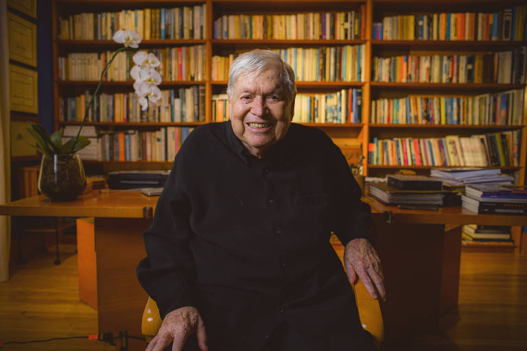 Um homem idoso, com cabelo grisalho e pele clara, sorri enquanto está sentado em uma cadeira. Ele usa uma camisa preta e está posicionado em frente a uma mesa de escritório. Ao fundo, há uma estante repleta de livros e uma planta com flores brancas. A iluminação é suave, destacando o rosto do homem e o ambiente acolhedor.