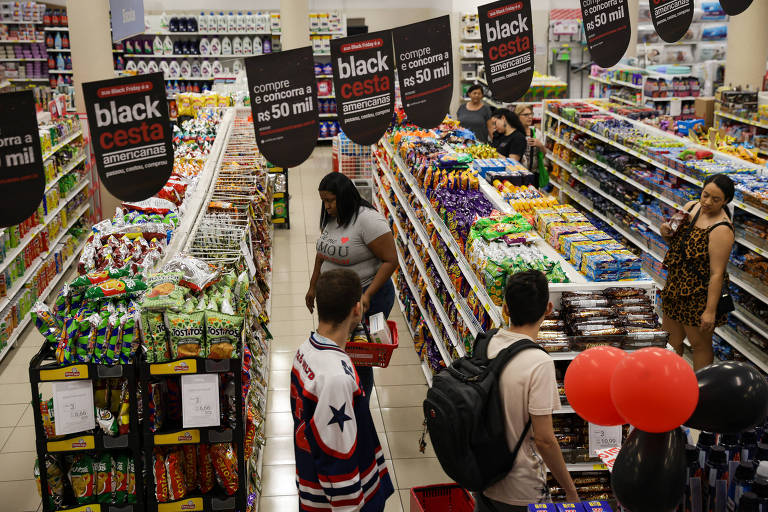 Imagem mostra o ambiente interno de uma loja da Americanas. Foto foi tirada do alto e mostra 3 corredores da loja com prateleiras de produtos e alguns clientes no local. Também aparecem bandeirinhas penduradas no alto com mensagens sobre a promoção da Black Friday