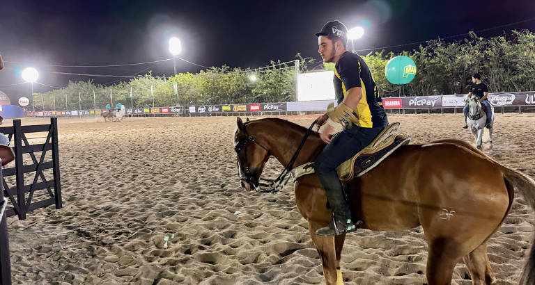 A imagem mostra um cavaleiro montado em um cavalo marrom em uma arena de areia iluminada por luzes artificiais. O cavaleiro usa uma camisa preta e amarela e um boné. Ao fundo， outros cavaleiros estão em ação， e há uma grande bola verde visível. O ambiente é de um evento equestre noturno.