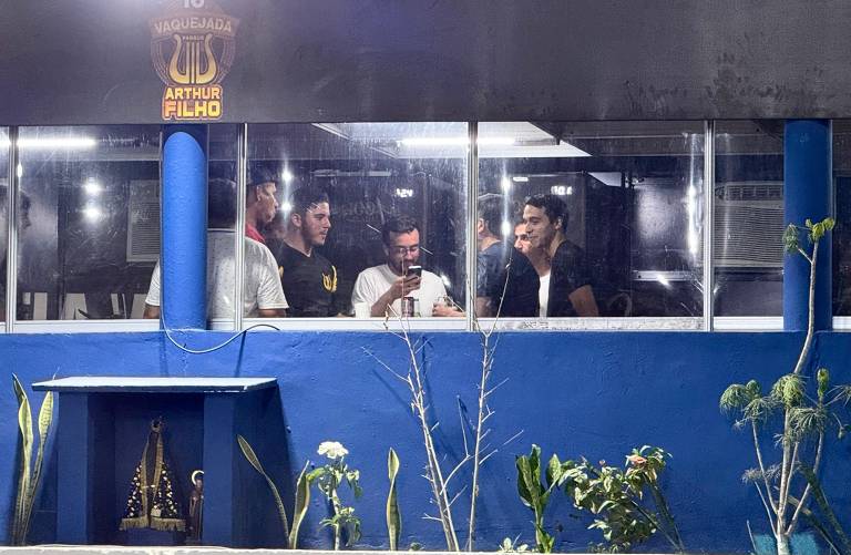 A imagem mostra um grupo de seis homens dentro de um ambiente com janelas de vidro. Eles estão em uma conversa animada, com um deles olhando para um celular. O ambiente é decorado com uma parede azul e há uma pequena área externa com plantas e uma imagem religiosa em um altar. O logotipo 'Vaquejada Arthur Filho' está visível na parte superior da imagem.