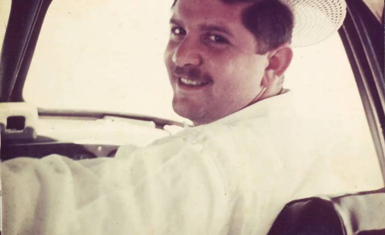 Homem branco de bigode curto sorri enquanto olha para trás. Ele veste uma camisa branca e tem chapéu na cabeça